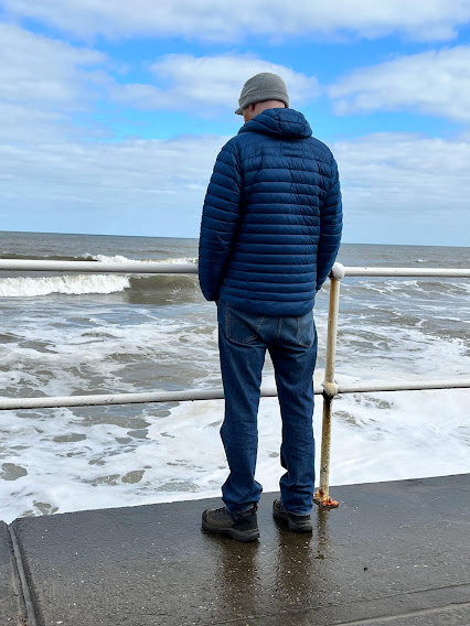 A man in a blue coat, jeans, and grey hat facing away from the camera watching the ocean over a white railing.